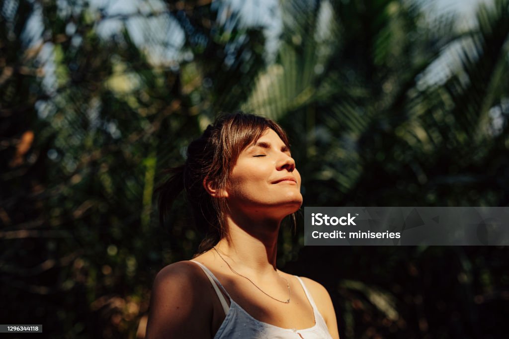 Beautiful Happy Woman Enjoying the Warm Sunlight in a Tropical Public Park Close up upper body shot of a beautiful happy young Caucasian woman enjoying the warm sunlight and tropical atmosphere with her eyes closed surrounded by palm trees in a tropical public park. Women Stock Photo