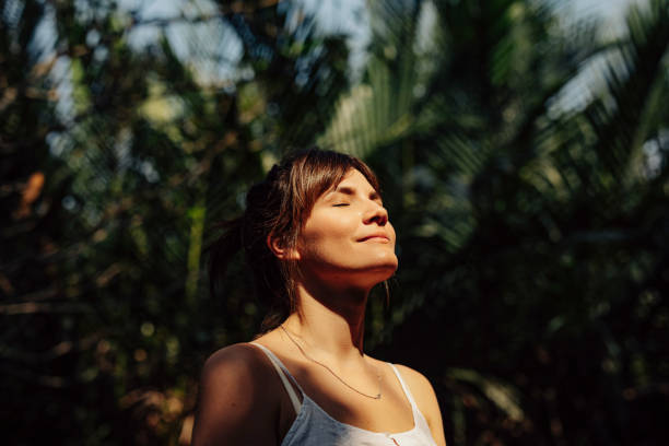 Beautiful Happy Woman Enjoying the Warm Sunlight in a Tropical Public Park Close up upper body shot of a beautiful happy young Caucasian woman enjoying the warm sunlight and tropical atmosphere with her eyes closed surrounded by palm trees in a tropical public park. the natural world stock pictures, royalty-free photos & images