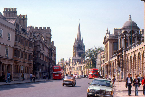 High Street, Oxford England, May 5 1983. The image includes some students, cars and red double decker busses with the iconic historic colleges in honey colored stone that line this main thoroughfare in the English university city. A normal day in Oxford back in 1983