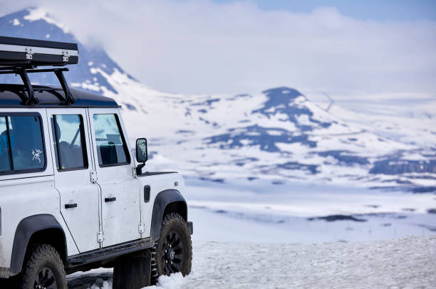 A white 4x4 landrover defender standing on a mountain. It ‘watches’ over the mountains in front of it that are covered in snow. The sky is mainly white because of the clouds.