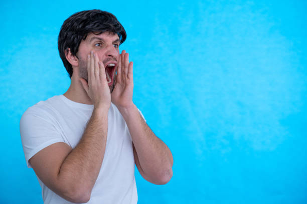 istock Man standing over blue background Shouting angry out loud with hands over mouth 1296350022