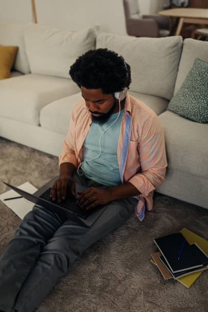 istock Young black man with earphones sitting on the floor and using laptop 1296358095