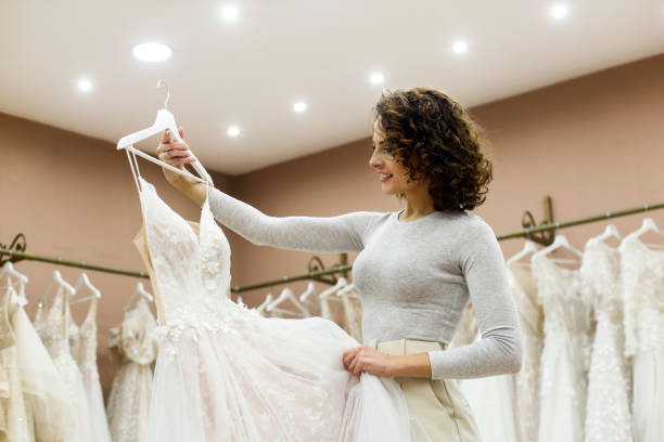 Young woman looking at a wedding dress in a bridal shop Young woman choosing a wedding dress in a bridal shop. About 25 years old, Caucasian brunette. bridal boutique stock pictures, royalty-free photos & images