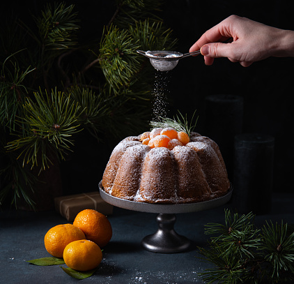 A woman's hand sprinkles powdered sugar on a tangerine Christmas cake. Dark and mood image