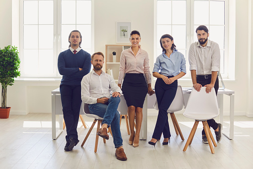 Full length group portrait of young business team in office. Start-up founder with partners looking at camera. Confident financial company chief executive surrounded by group of happy loyal employees