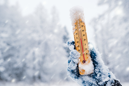 Human hand holding a thermometer with the low temperature against winter nature background with snowfall.