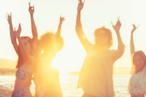 Group of friends dancing and celebrating on the beach at sunrise/sunset. They are very happy, smiling and laughing. Some have their arms raised. The sea and sun can be seen in the background. Defocussed with focus on background