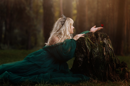 A beautiful blonde young woman in a long green dress and a diadem on her head in the forest. girl sitting near the old stump with butterfly on her hand. Solar glare. Fantasy. fairy tale.