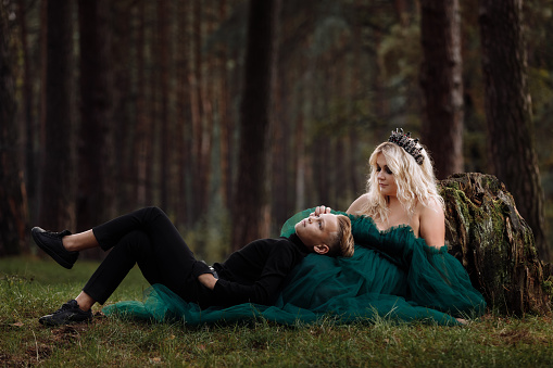 A beautiful blonde young woman in a long green dress and a diadem on her head with stylish young son in the forest. girl and boy sitting near the old stump. Solar glare. Fantasy. fairy tale.