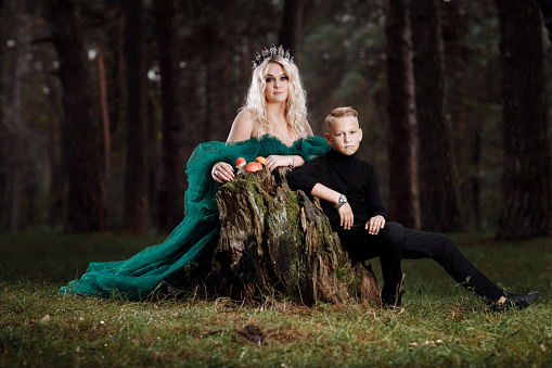 A beautiful blonde young woman in a long green dress and a diadem on her head with son in the forest. girl and boy sitting near the old stump with amanitas. Solar glare. Fantasy. fairy tale.