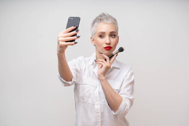 verticale de studio de belle fille à la mode avec le cheveu court et composent le bras de fixation sur la taille et prenant le selfie par l’intermédiaire du téléphone portable au-dessus du fond d’isolement blanc - stage makeup white caucasian fashion photos et images de collection