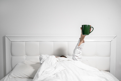 Woman hand lifts up green mug of drink from under soft blanket lying in large comfortable bed with pillows in light bedroom