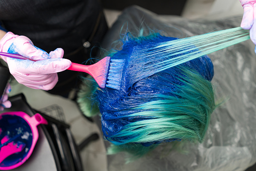 Top view of hair coloring in beauty salon. Hairdresser in protective glove using pink brush while applying blue paint to customer with emerald hair color during process of dyeing hair in unique color