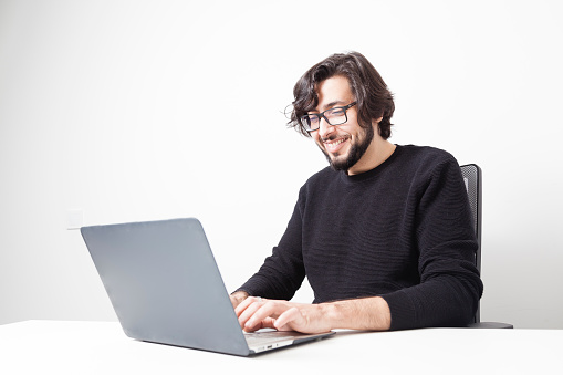 Middle eastern adult bearded man with long hair and glasses using laptop computer while smiles on a white office room with copy space.