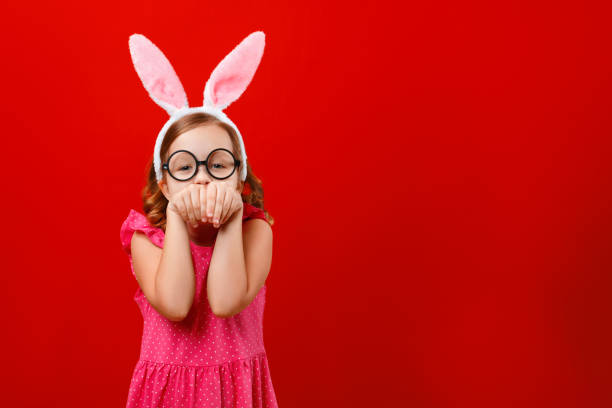 幸せなイースター。赤い背景にバニーの耳と眼鏡で陽気な小さな女の子の肖像画。子供は楽しみを持っており、ウサギのふりをします。 - easter easter bunny fun humor ストックフォトと画像