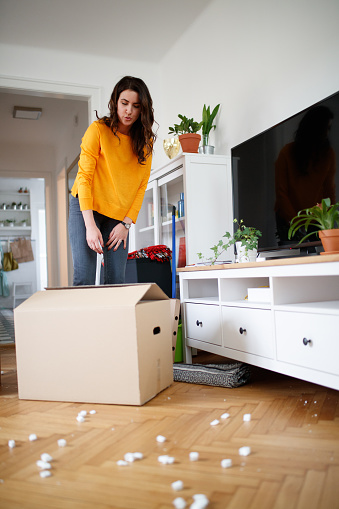 Woman standing in living room and packing for house move, cardboard box in front of her