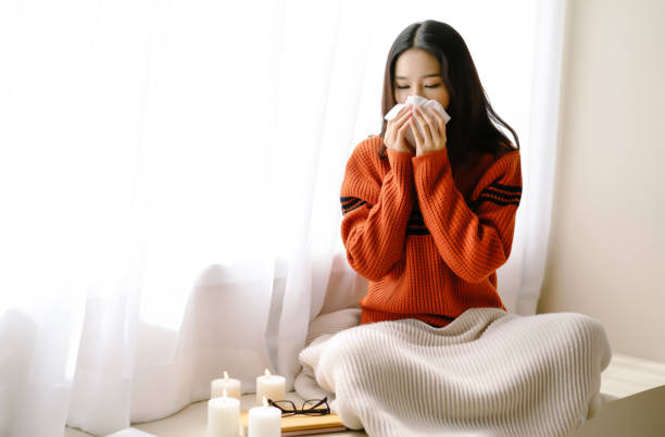 retrato de la joven hermosa mujer asiática estornudando y soplando la nariz con un pañuelo sentado en casa en la silla junto a la ventana. la joven asiática enferma de gripe en un día de invierno. mujer con un resfriado. - sniffles fotografías e imágenes de stock