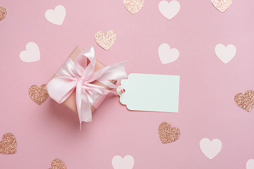 Gift box with blank gift tag on pastel pink background with many hearts. Valentines day composition. Top view, flat lay.