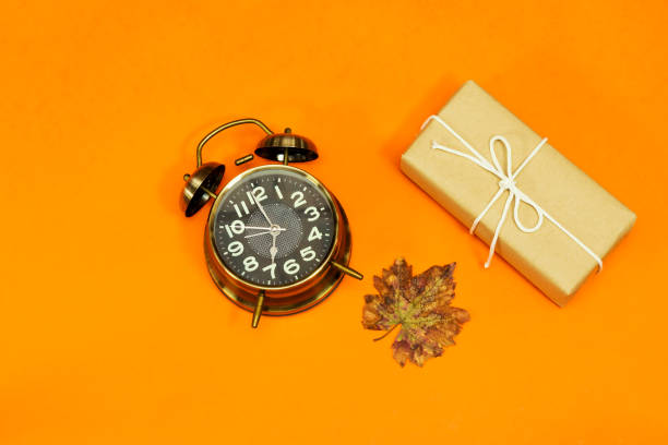 alarm o'clock showing 7 o'clock and autumn leaves, gift box isolated on orange background. time to holidays - clock time alarm clock orange imagens e fotografias de stock