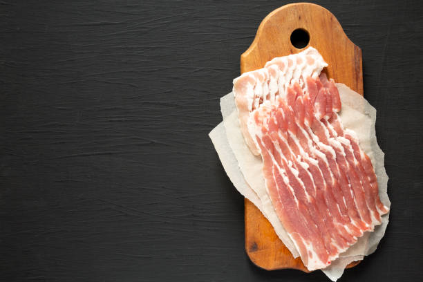 istock Uncooked Raw Bacon on a rustic wooden board on a black surface, overhead view. Flat lay, from above, top view. Copy space. 1296464907