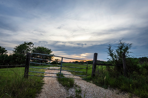 Scenic landscape of a gravel lane leading into a pasture blocked by a weathered, crooked metal gate with the sun setting while a storm is blowing in.