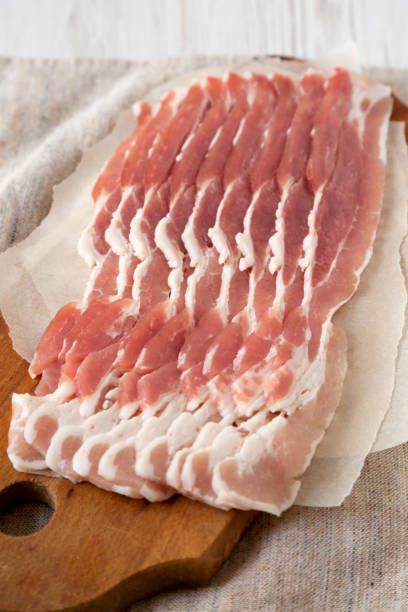 istock Uncooked Raw Bacon on a rustic wooden board on a white wooden background, side view. Close-up. 1296465908
