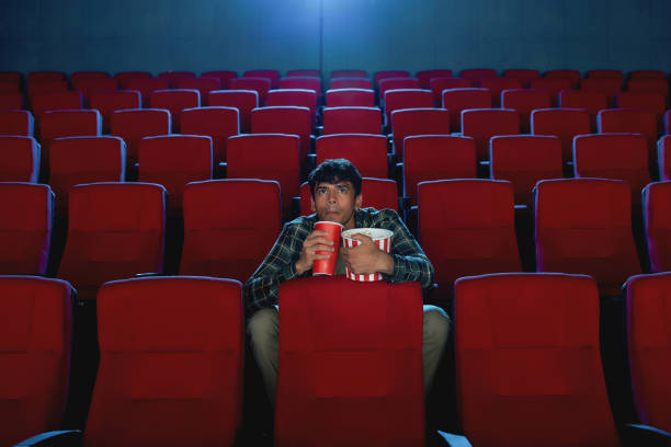 istock Focused young man holding a drink and popcorn basket while watching movie alone in empty theater auditorium 1296473243