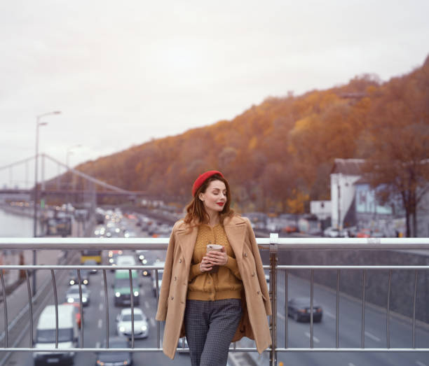 istock Charming Parisian young woman drinks coffee using reusable mug looking at camera smiling standing at pedestrian bridge with cars on the road. Looking happy fashion girl in a red beret and beige coat 1296475785