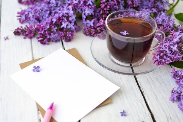 Delicate morning tea table setting with lilac flowers, a transparent cup of tea and a saucer and a white vase, white and craft paper for notes, and a pink pen on a white wooden table. Mockup. Copy space. Spring breakfast concept. delicado ajuste de la mesa de té de la mañana con flores lilas, una taza de té transparente y un platillo y un jarrón blanco, papel blanco y artesanal para notas, y un bolígrafo rosa sobre una mesa de madera blanca. maqueta. copiar espacio. concepto d