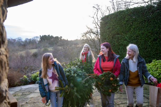Lets Get The Tree Indoors Front view of a mother with her adult daughter and her children as they walk into the family home carrying the Christmas tree which they've just bought one of the daughters is also carrying a Christmas wreath the grandmother carrying the decorations for the tree in a paper bag. 12 17 months stock pictures, royalty-free photos & images