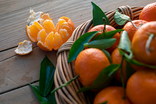 Close-up of a group of satsumas with leaves in a wicker basket on a wooden bench. There is one satsuma out of the basket on the bench, peeled and open.