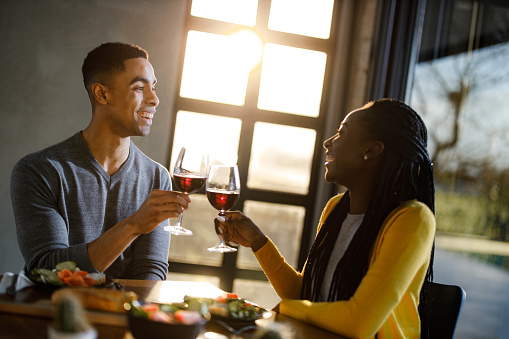 Happy African American couple communicating while toasting with wine during lunch at dining table. Focus is on man.