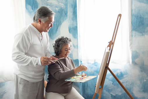 Asian Elderly Couple painting on canvas at their home. Happy Smiling Old man and woman colouring together.