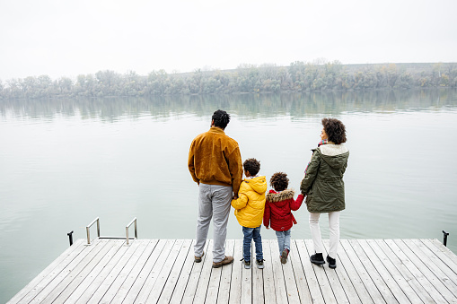 Back view of African American family holding hands on a jetty and looking at river view in winter day. Copy space.