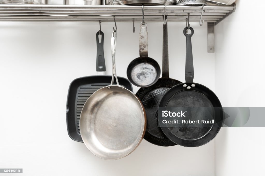 Various pans in different sizes and forms for cooking and frying hanging on metal hooks from shelf in kitchen with white wall in background Cooking Pan Stock Photo