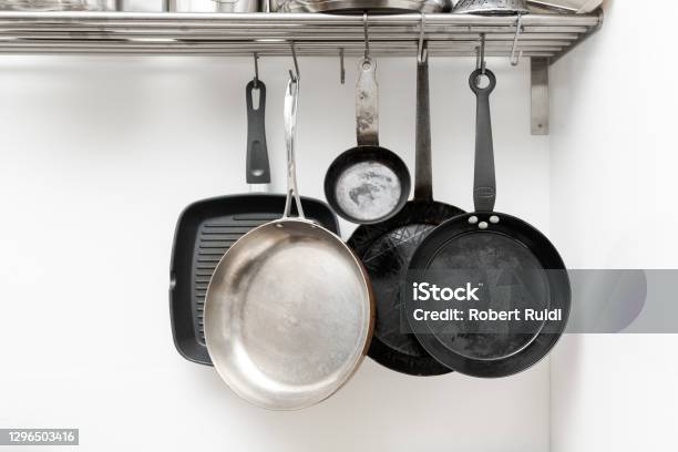 Various Pans In Different Sizes And Forms For Cooking And Frying Hanging On Metal Hooks From Shelf In Kitchen With White Wall In Background Stock Photo - Download Image Now