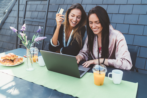 Young women having breakfast at terrace and shopping online on laptop
