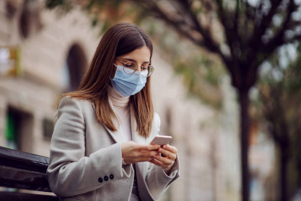 istock Young brunette with face mask on face sitting on the bench outside and using smart phone for texting. Millennials during corona virus. 1296512930