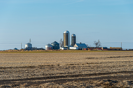 Bare ground winter farm field with farm silos, buildings and equipment in the background.