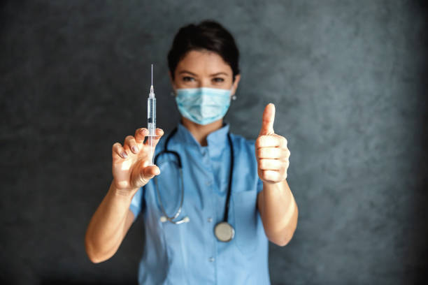 doctor with mask on face holding covid-19 vaccine and shoving thumbs up. vaccine is tested and ready for use. - injecting flu virus cold and flu doctors office imagens e fotografias de stock