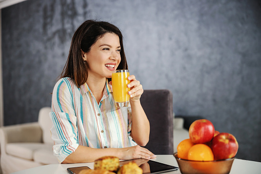 Smiling woman sitting at dining table in the morning and having healthy breakfast. Woman drinking orange juice.