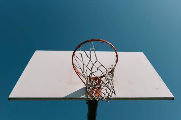 istock Looking up at old basketball hoop with torn net and white backboard alone against blue sky. 1296517849