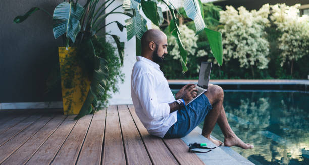 vista lateral de un experto hombre de negocios que utiliza el dispositivo portátil para hacer entradas en línea y la banca durante la recreación del complejo en la isla de bali, nómada digital de oriente medio que se conecta a la villa wifi en netbook - teletrabajo fotografías e imágenes de stock