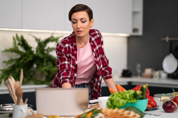 istock Young woman cooking in the kitchen. Using a laptop computer while cooking young housewife searching online recipes or watching online tv show while cooking. Healthy life living 1296530158