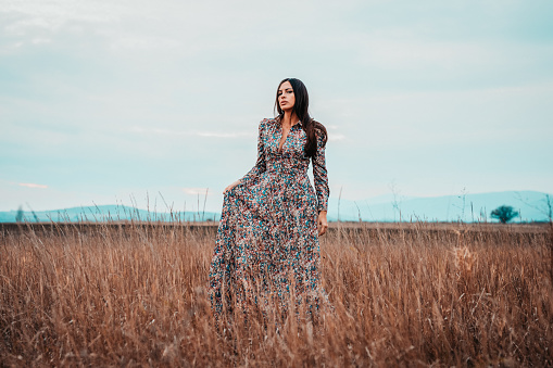 Portrait of a beautiful young woman with long brown hair wearing longsleeved floral pattern dress in nature on a lovely autumn day