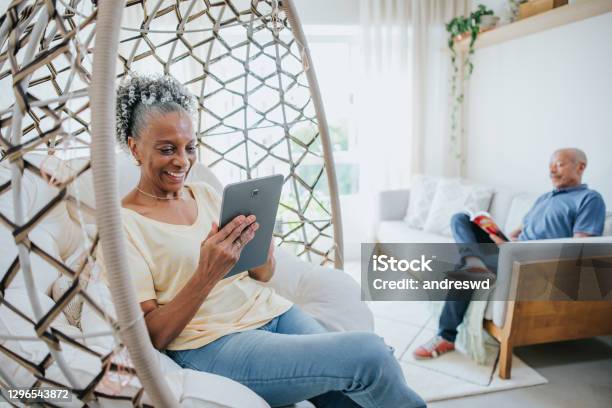 Senior Couple In Living Room Man Reading Book And Woman Using Digital Tablet Stock Photo - Download Image Now