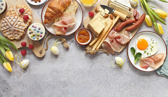 Easter Breakfast food table. Festive brunch set, meal variety with fried egg, croissant sandwich, cheese platter and desserts. Overhead view