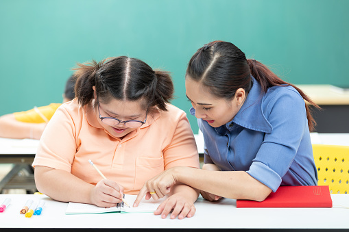 portrait asian disabled child or autism child writing a book and woman teacher helping in classroom