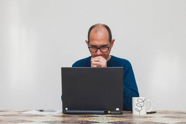 A man wearing glasses and a blue jersey is pensive in front of a laptop. On the table there is a white cup, a smartphone, a sheet of paper and a pen.