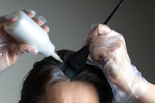 Cierra las manos de la mujer teñiendo el cabello usando cepillo negro. Mujer de mediana edad coloreando el pelo oscuro con raíces grises en casa photo